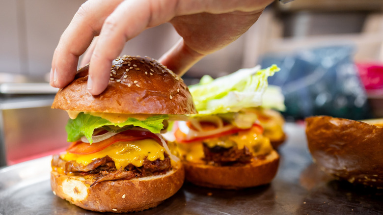 A hand placing down the top bun of a burger with more burgers int he background