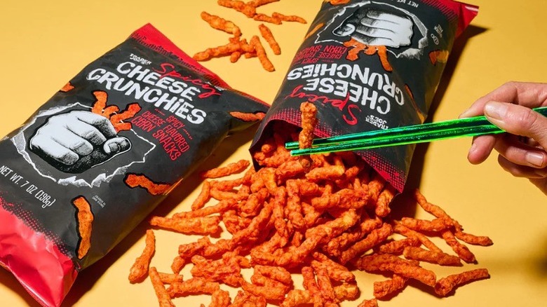 two bags of Trader Joe's Spicy Cheese Crunchies pictured with a hand holding one piece of the snack in a pair of green chopsticks