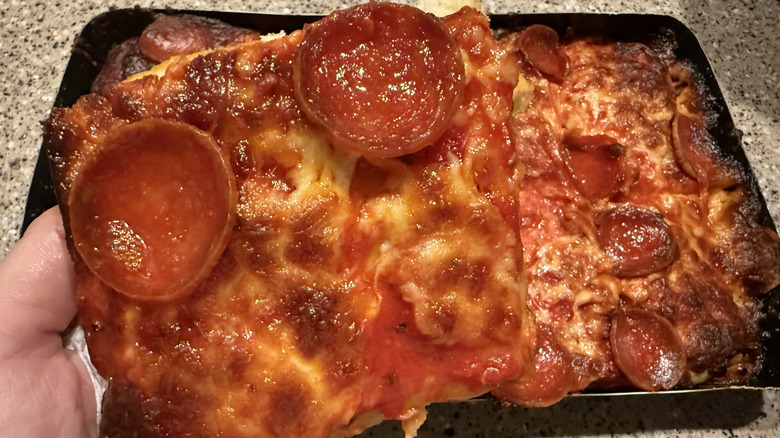 Close up of slice of cooked Trader Joe's Detroit Style Pepperoni Pizza above rest of pizza in tray on granite counter