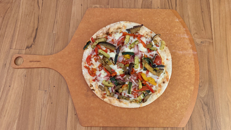 A thin crust pizza piled with vegetables on a pizza peel set on a wooden table.