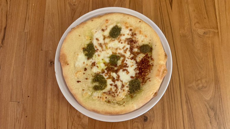 A small round pizza with six dollops of green pesto and brown roasted garlic on a white cheese.