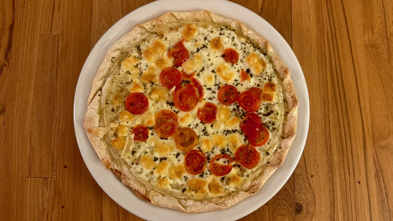 A thin pizza with a braided crust that is dotted with bright red halves of cherry tomato.