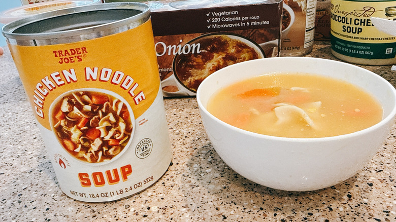 Can of Trader Joe's Chicken Noodle Soup next to some in a white bowl with other TJ's soups in background