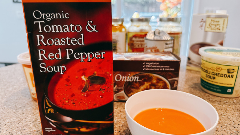 Trader Joe's Organic Tomato and Roasted Red Pepper soup next to some in a white bowl with other TJ's soups in background