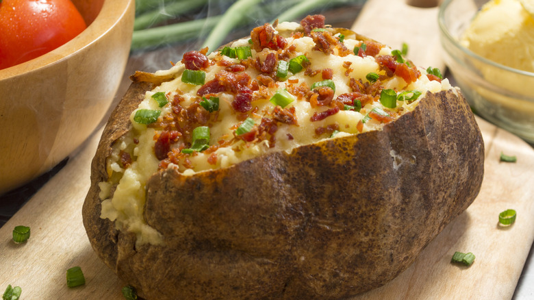 A large baked potato topped with chives and bacon, sitting on a wooden cutting board