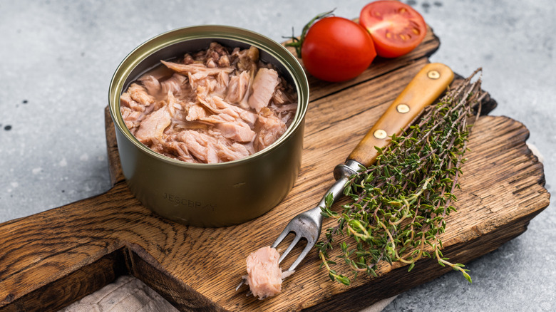 An opened can of tuna on a wooden cutting board beside cherry tomatoes and herbs