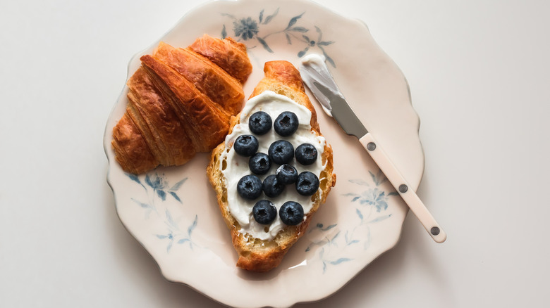 Croissant sliced win half on plate with one slice spread with yogurt and topped with fresh blueberries