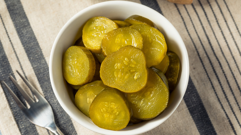 A white bowl of bread and butter pickle chips and fork on a cloth surface.