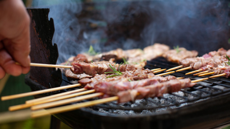 A hand adds a meat skewer to a row of similar skewers over a charcoal grill
