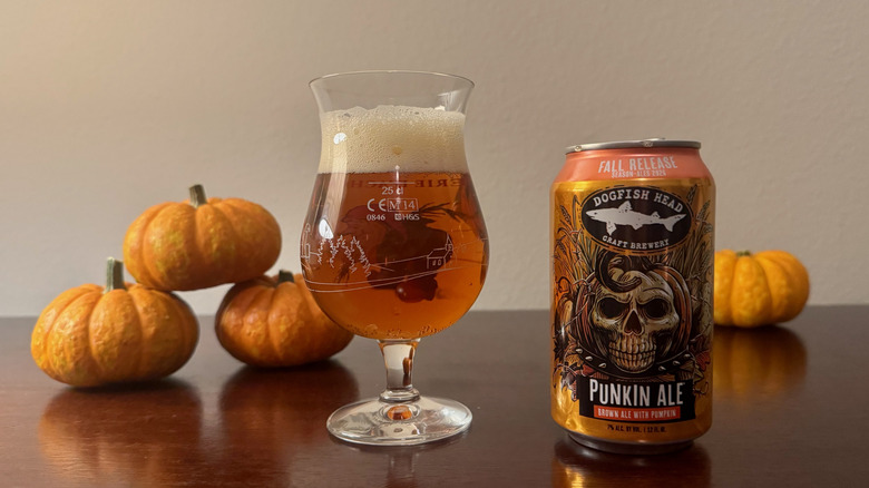 A full glass of pale amber beer next to a can of Dogfish Head Punkin Ale with a smiling skull logo.