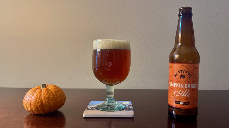 A glass beer bottle  of Kentucky Pumpkin Barrel Ale with an orange label next to a dark amber beer and a small pumpkin.
