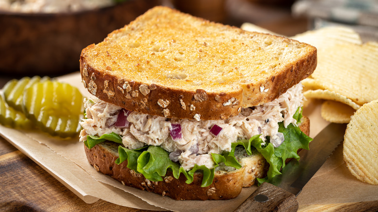 Tuna salad sandwich on sliced wheat bread alongside potato chips and pickles