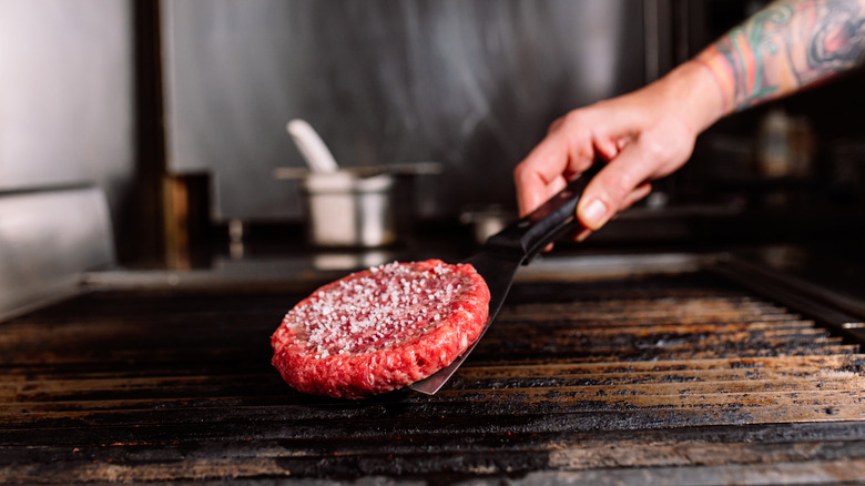 A hand placing a salted burger patty on a grill with a metal spatula