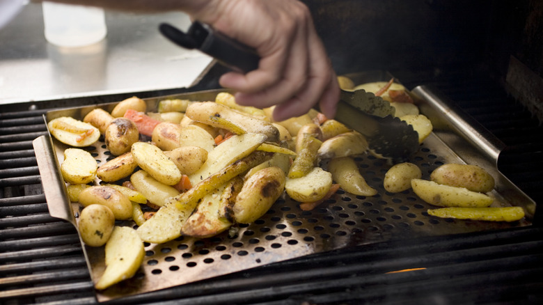 Someone grilling fingerling potatoes and turning them with tongs on a large outdoor gas grill.