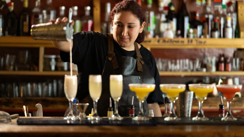 Adult woman bartender pours drink from metal cocktail shaker into a glass to add to a line of colorful mixed drinks