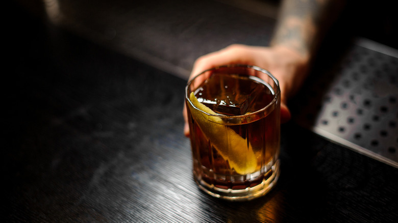 Man serving a glass of a Rusty Nail cocktail with orange zest on the bar counter