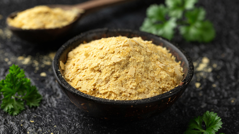 Small black bowl filled with nutritional yeast flakes with wooden spoon in the background also filled with nutritional yeast