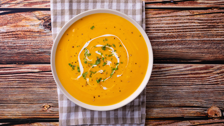 A bowl of pumpkin soup rests on a tablecloth atop a wooden table