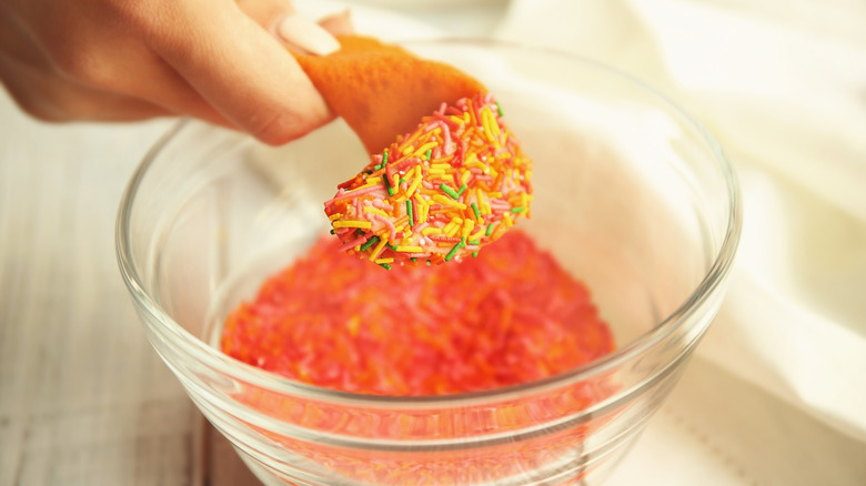 A woman dips a fortune cookie into a bowl of colorful sprinkles