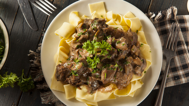 A white plate with noodles topped with beef stroganoff sits on a wooden table with a chequered napking and cutlery at the side