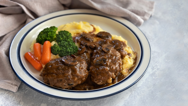 Close up of plate with Salisbury Steak, mushrooms, mashed potato, broccoli, carrots and gravy on gray background.