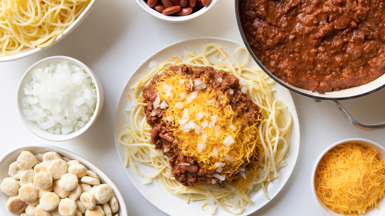 Bowl of Cincinnati chili with pasta next to bowls of cheese, onions, and chili