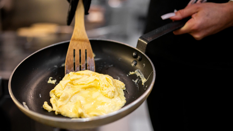 A gloved hand using a wooden spatula to cook fluffy scrambled eggs as another hand holds the non-stick frying pan over the flame.
