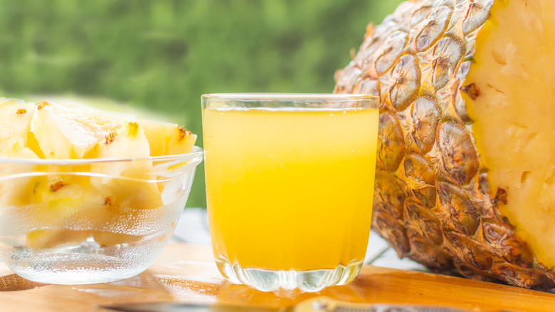 A small glass filled with pineapple juice beside cut up pieces of pineapple in a bowl and a large pineapple cut in half