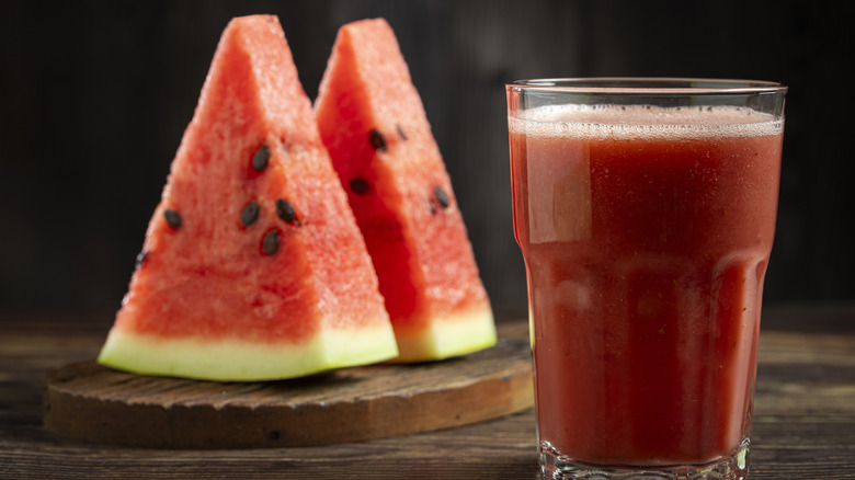 Two slices of watermelon stood up on a wooden board beside a glass of fresh watermelon juice