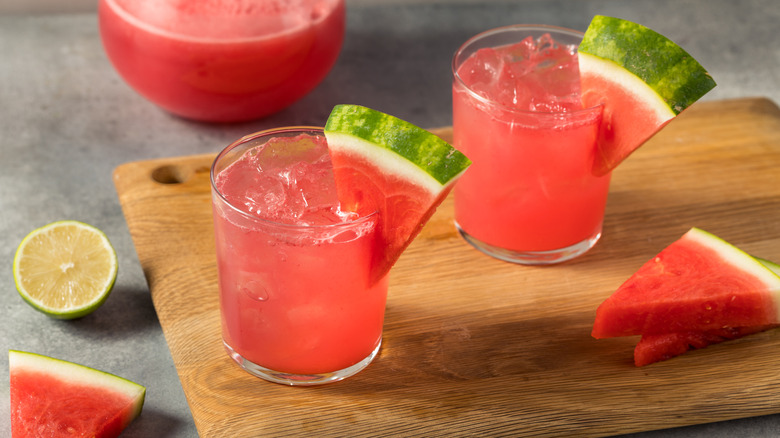 Two glasses of a watermelon cocktail with watermelon slices on the rims on a cutting board with a lime beside it