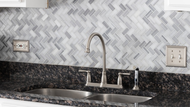 Zigzag backsplash in shades of gray behind a metal kitchen sink in a marbled black tile counter