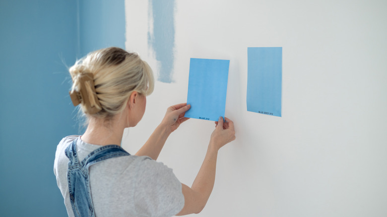 Woman holding square blue paint sample with another one a white wall in front of her that's partially painted a turquoise blue