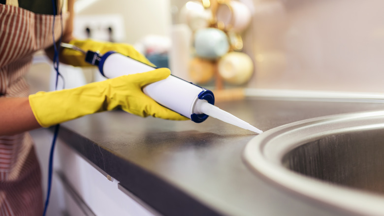 Close-up of woman wearing yellow gloves using a caulk gun on a metal kitchen sink in a black counter