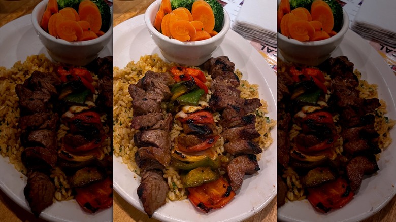 Texas Roadhouse steak kabobs on white plate over rice with side dish of carrots and broccoli