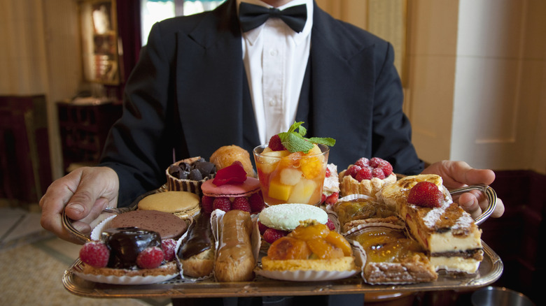 A waiter in tuxedo serves a tray full of fancy desserts