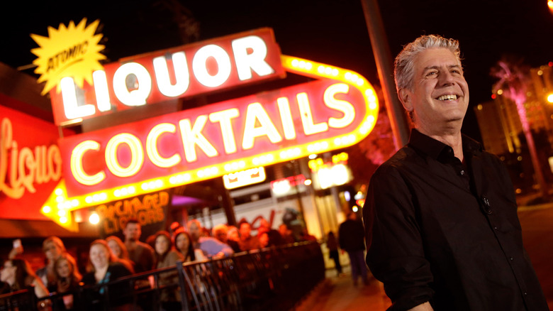Anthony Bourdain smiling in front of a large neon sign that reads "Atomic Liquor Cocktails"