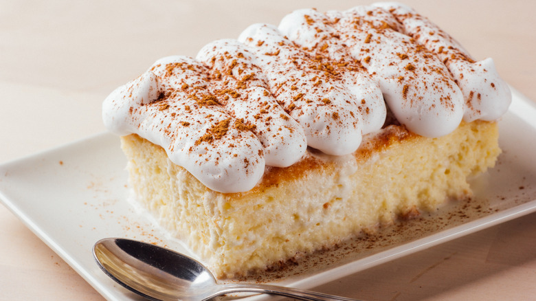 A square of tres leches cake topped with whipped cream on a white plate