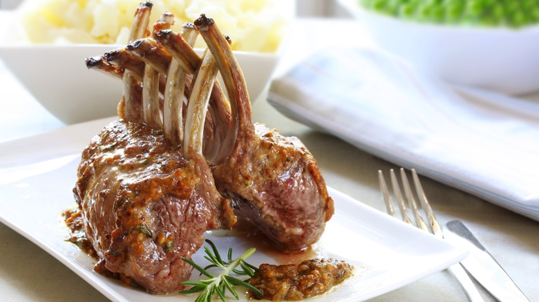 Roast rack of lamb on a white plate with silverware and a bowl of mashed potatoes