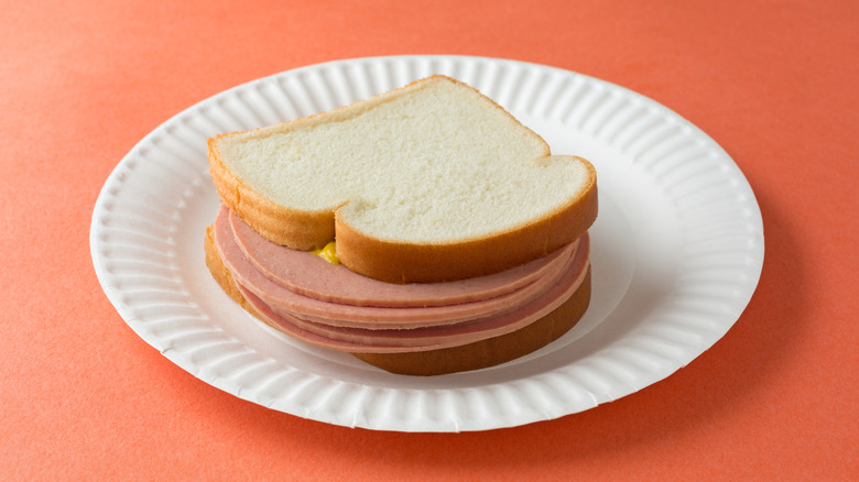 A bologna sandwich with mustard on white bread on a white paper plate