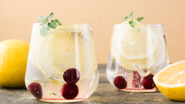 Two short glasses with hard seltzer, lemon and cherries in the glass with garnish at the top, all on a dark, texturized table.