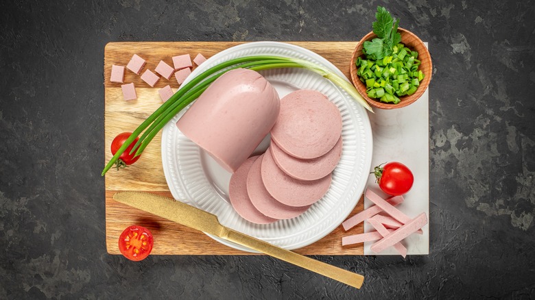 Sliced bologna next to a bologna loaf, surrounded by veggies on a cutting board