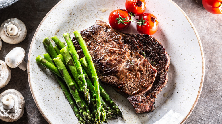 A well-done steak on a white plate with asparagus and cherry tomatoes