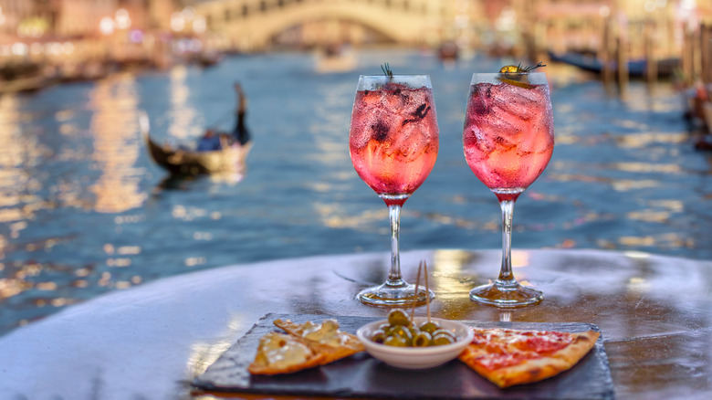 Two slices of pizza plated on a table with pink drinks in wine glasses, in front of a gondola on a canal
