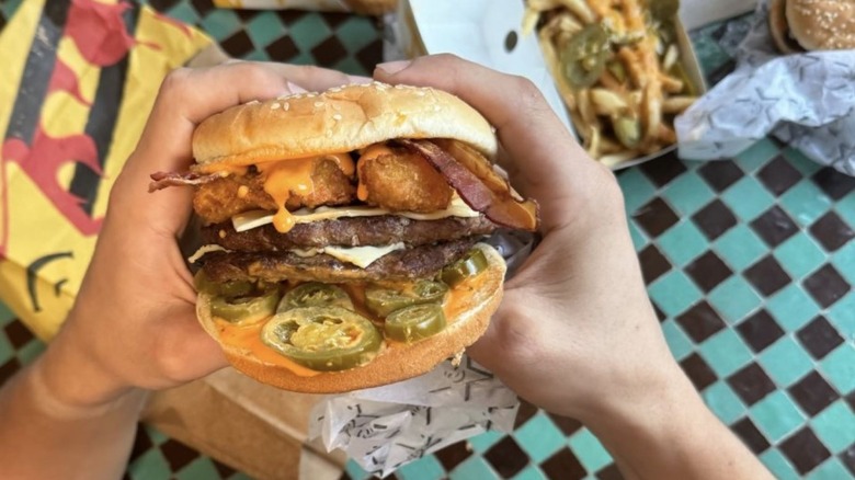 Two hands holding a burger with two patties, cheese, jalapeno slices, bacon, and fried jalapeno poppers with an orange sauce