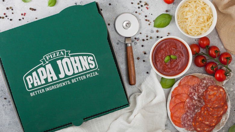 A green pizza box with Papa John's logo on it next to bowls and plates of pizza ingredients like tomatoes, cheese, meat, and sauce.