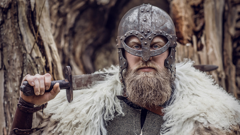 A man dressed in Viking garb including a helmet and fur coat with a sword resting on his shoulder.
