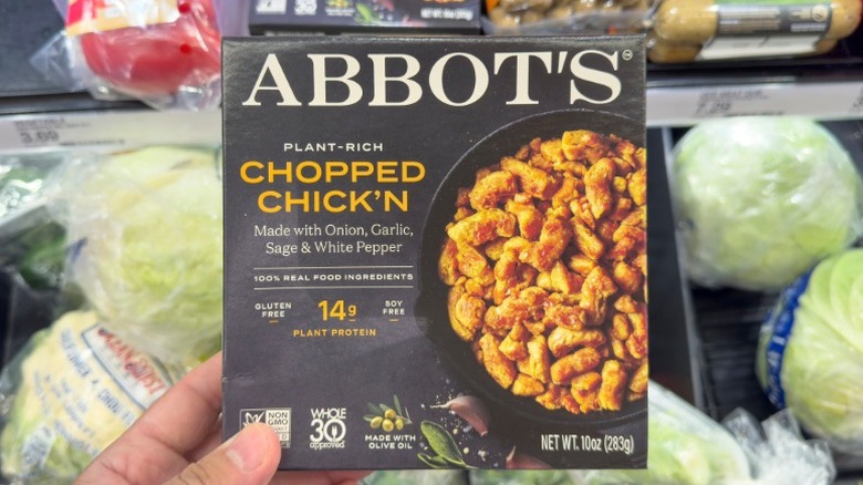 A hand holding a package of Abbot's plant-based chopped chicken in the produce aisle of a grocery store