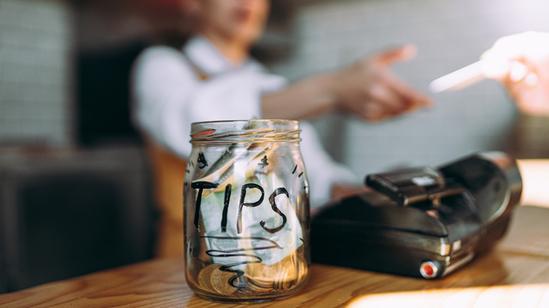 A counter with a tip jar on it while a worker reaches for a bank card in the background