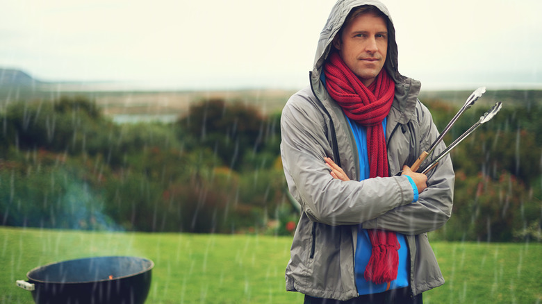 Man standing in the rain holding tongs while it's raining with a smoking grill in the background
