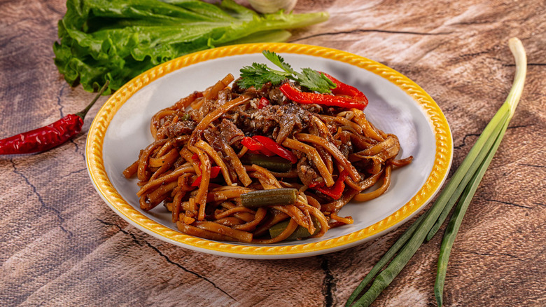A stir-fry with beef, noodles, peppers, and green onions in a shallow white bowl with a yellow rim.